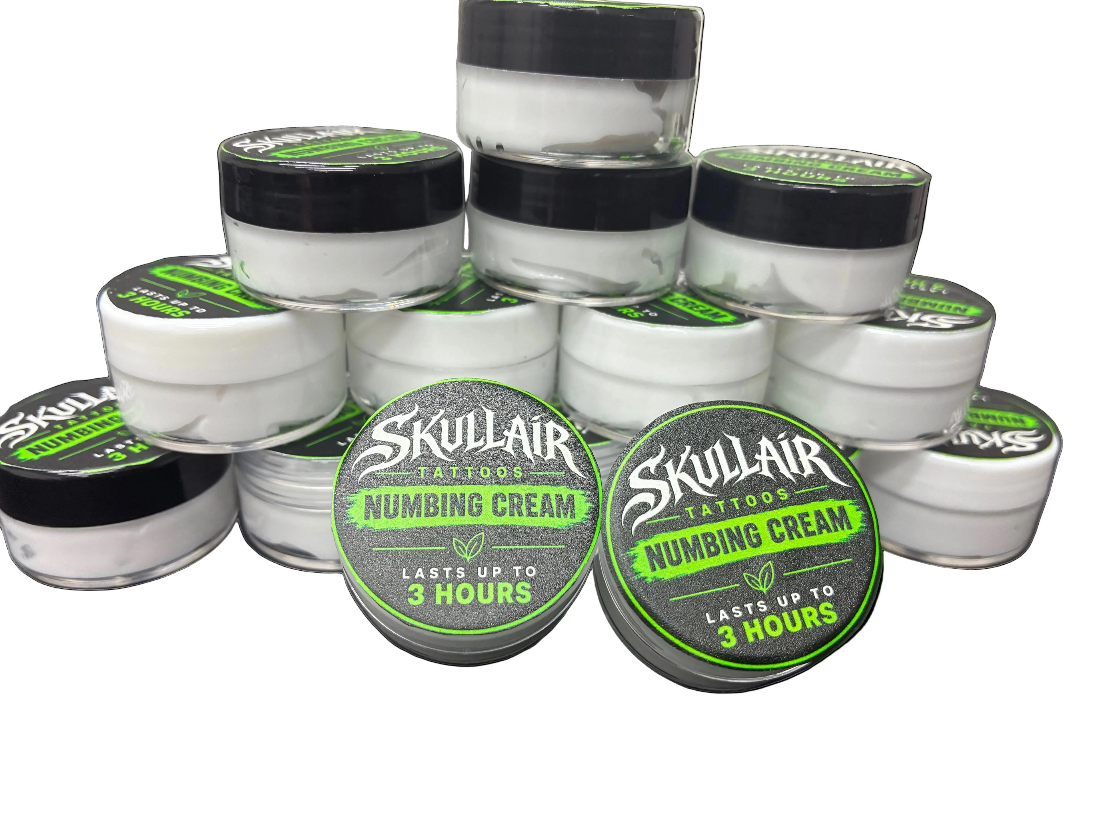 A stack of small white plastic jars of Skullair Tattoos Numbing Cream with black lids, arranged on a dark textured surface. Two jars in the front have green and black labels showing the brand name and product details.