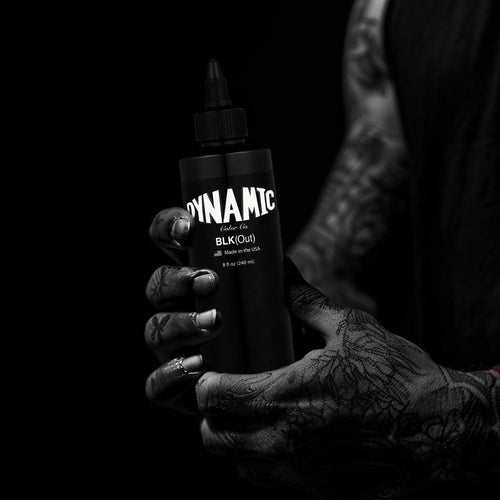 Tattooed hand holding a Dynamic Blackout tattoo ink bottle against a dark background